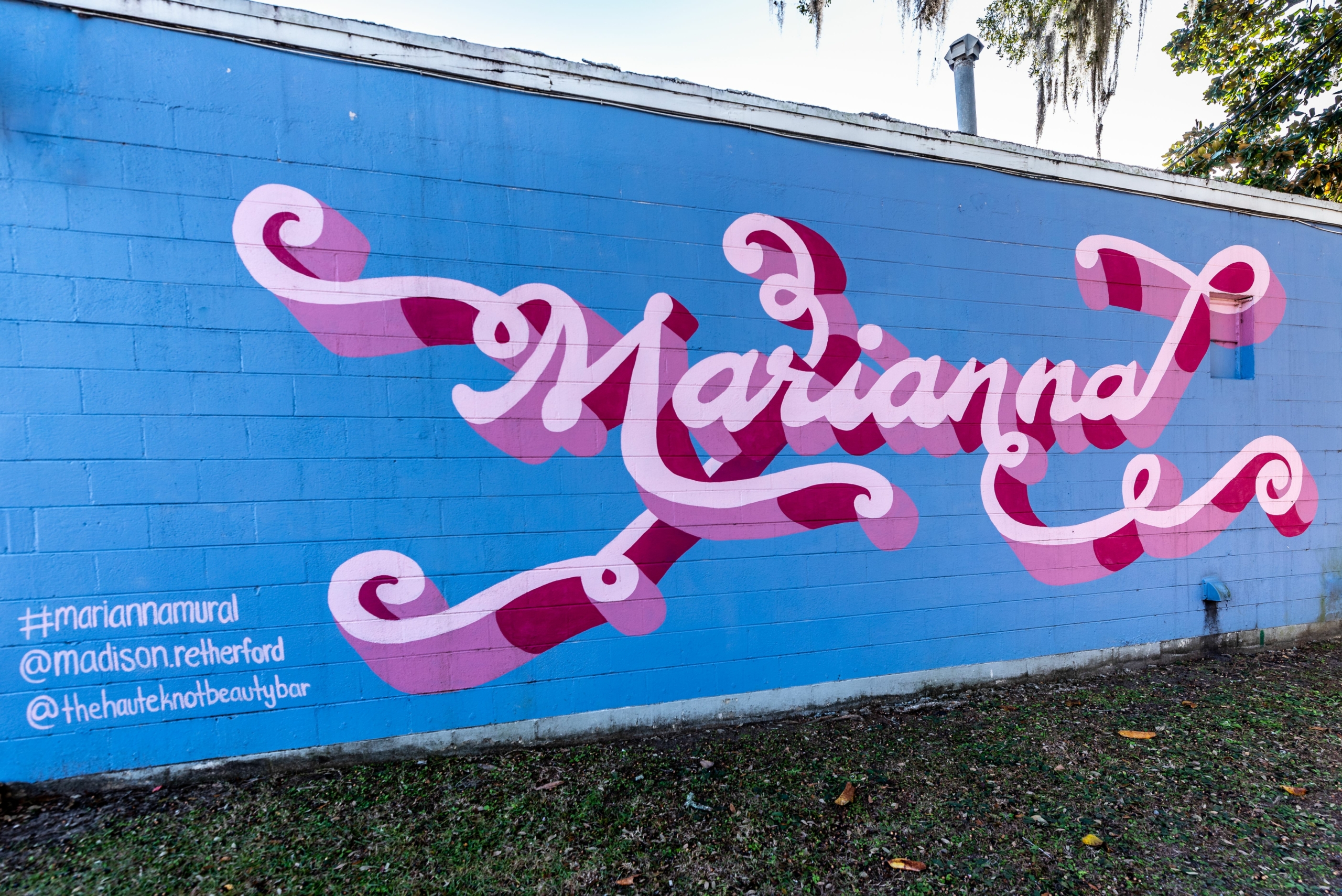 A wall mural with blue background with Marianna painted in curly script in shades of pink.