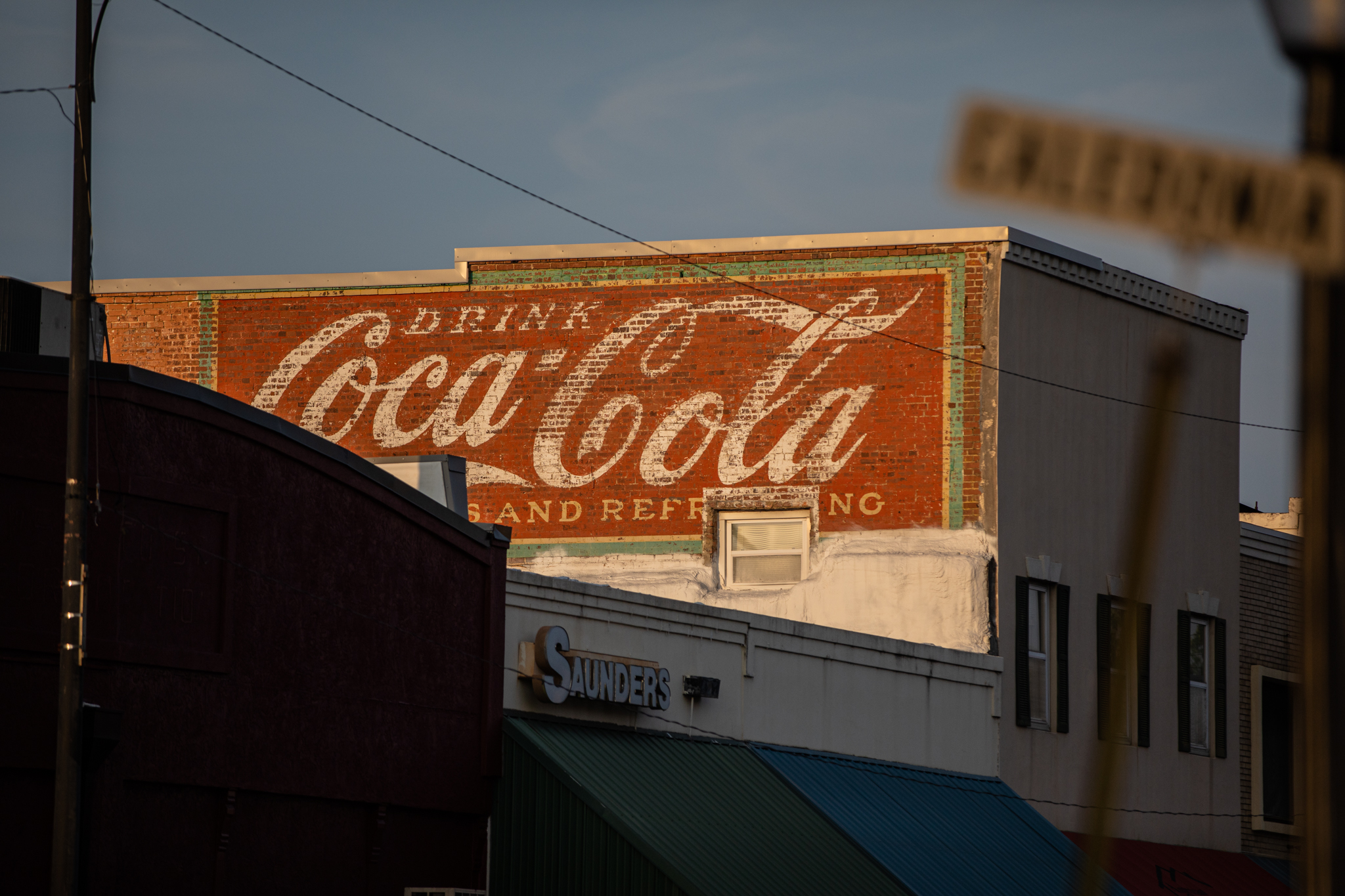 The sun sets on an old Coca-Cola mural on the side of a building
