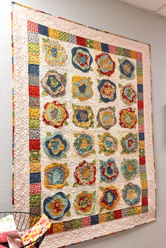 Brightly colored ruffled French rose quilt with block border hangs on the wall.