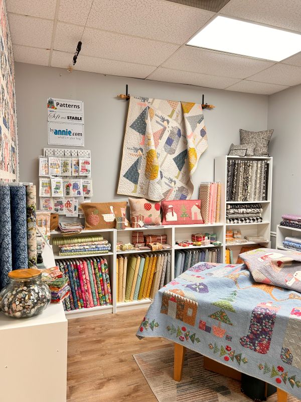 A cozy quilting shop interior with colorful quilts displayed on the wall and bed, fabric bolts neatly organized on white shelves, and a variety of sewing notions and handmade pillows. A must stop for Florida Shop Hop.
