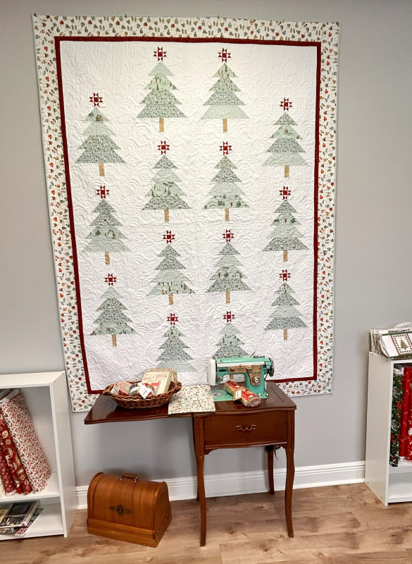 A Christmas tree quilt hangs on the wall behind a vintage teal sewing machine with vintage sewing table ready for Christmas in July.